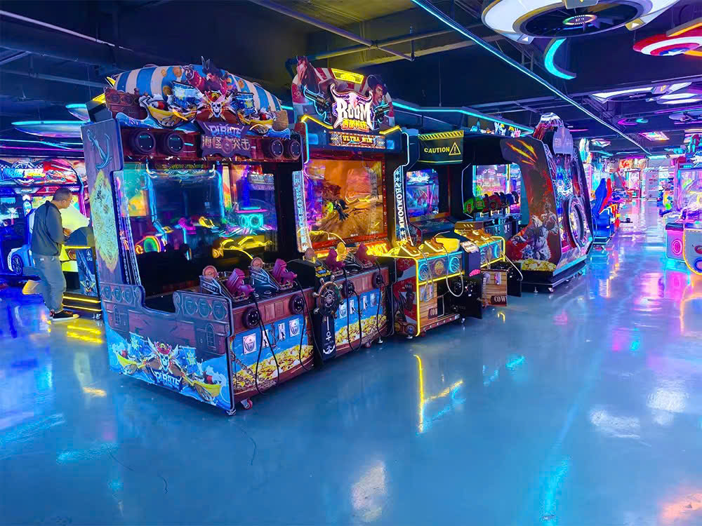 Khu vực phòng chơi với nhiều máy game chủ đề hải tặc xếp hàng, ánh sáng neon xanh lam chiếu sáng sàn và tủ máy, người chơi đang trải nghiệm Khu vực phòng chơi với nhiều máy game chủ đề hải tặc xếp hàng, ánh sáng neon xanh lam chiếu sáng sàn và tủ máy, người chơi đang trải nghiệm