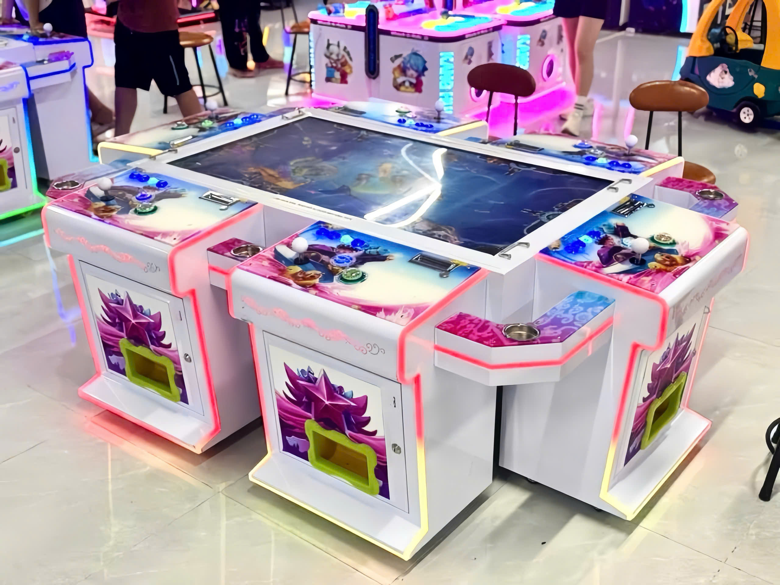 Máy bắn cá 6 người chơi bàn lớn arcade tại khu vui chơi, đèn LED neon xanh hồng, xung quanh có xe điện trẻ em và trò chơi khác Máy bắn cá 6 người chơi bàn lớn arcade tại khu vui chơi, đèn LED neon vàng hồng