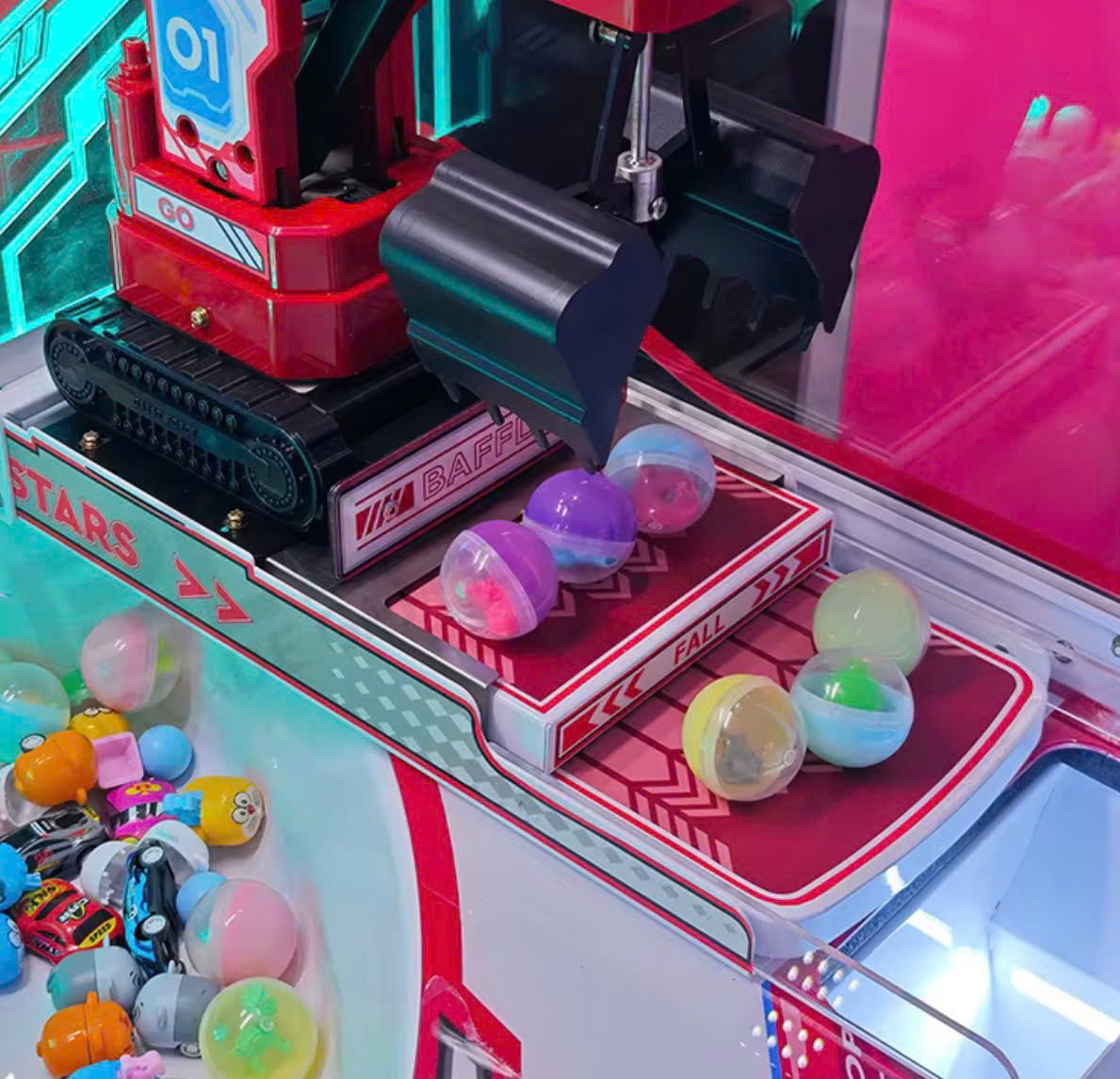 Cận cảnh càng gắp quà và cơ chế thả quà bên trong máy, với các quả bóng nhựa trên đường ray. Cận cảnh càng gắp quà và cơ chế thả quà bên trong máy, với các quả bóng nhựa trên đường ray.